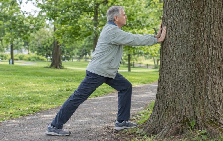 A man in his sixties doing a calf stretch against a park tree before a walk