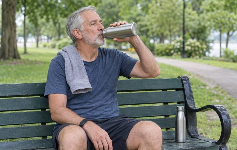 A man in his sixties drinking from a plain water bottle on a park bench after a walk