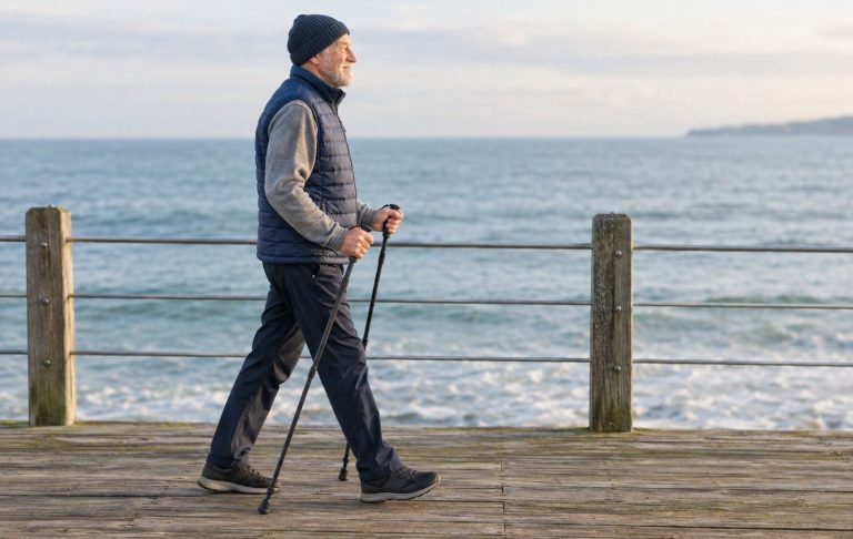 An older man walking on a seaside boardwalk with a pair of walking poles
